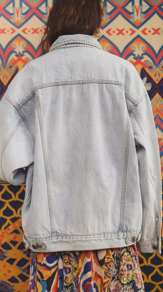 A photo of a denim jacket with a light wash. The jacket is worn by a woman with a colorful patterned dress. The background is a vibrant, patterned wall.