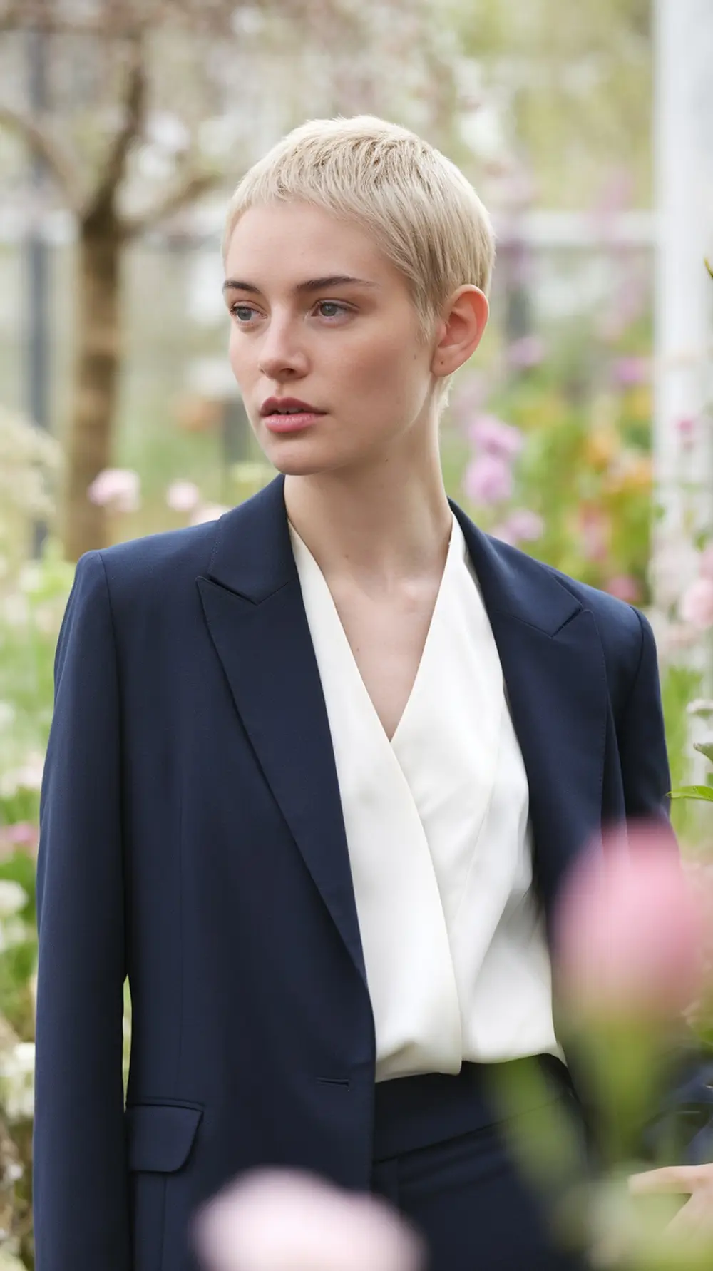 Model with a fresh pixie cut in a garden setting, wearing a blazer and a light top.