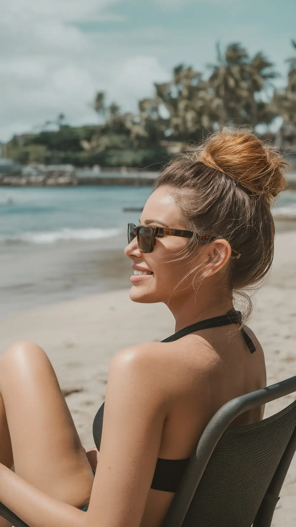 A woman with a high bun hairstyle sitting on the beach, wearing sunglasses.