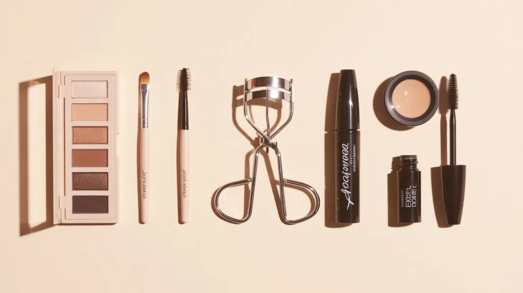 A clean flat lay photograph showcasing essential makeup tools for hooded eyes arranged on a soft cream-colored background. The composition features a neutral matte eyeshadow palette with warm brown and taupe shades, positioned alongside a small tapered blending brush, a precise pencil brush, a sleek eyelash curler, a tube of waterproof black mascara, and a compact gel eyeliner pot with its accompanying fine-angled brush. Each item is thoughtfully spaced with gentle shadows, creating an organized and elegant grid that feels both educational and approachable. The lighting is bright and even, eliminating harsh shadows while maintaining the clean, minimalist aesthetic that emphasizes the functionality and accessibility of each beauty tool.