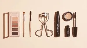 A clean flat lay photograph showcasing essential makeup tools for hooded eyes arranged on a soft cream-colored background. The composition features a neutral matte eyeshadow palette with warm brown and taupe shades, positioned alongside a small tapered blending brush, a precise pencil brush, a sleek eyelash curler, a tube of waterproof black mascara, and a compact gel eyeliner pot with its accompanying fine-angled brush. Each item is thoughtfully spaced with gentle shadows, creating an organized and elegant grid that feels both educational and approachable. The lighting is bright and even, eliminating harsh shadows while maintaining the clean, minimalist aesthetic that emphasizes the functionality and accessibility of each beauty tool.