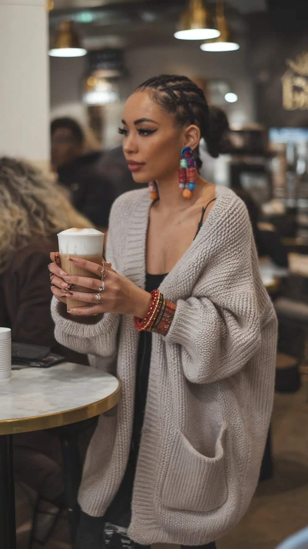 A woman in an oversized cardigan holding a latte, accessorized with colorful earrings and bracelets.